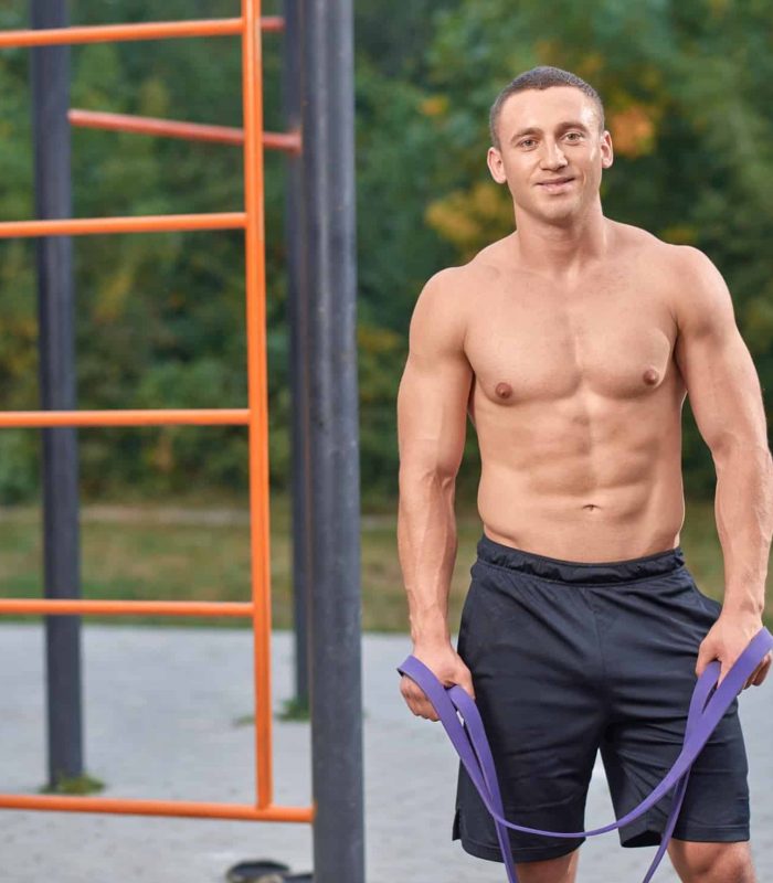 Handsome shirtless sportsman holding resistance, latex bands in hands. Attractive man posing during street workout. Muscular, athlete man standing, looking at camera, smiling.